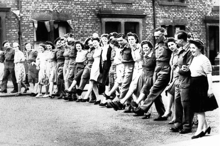 Men and women link arms across the street. They are all smiling and appear to be doing a jig or dance. Some of the men are dressed in uniform. There is a row of terraced houses behind them with bunting over the doors and windows.