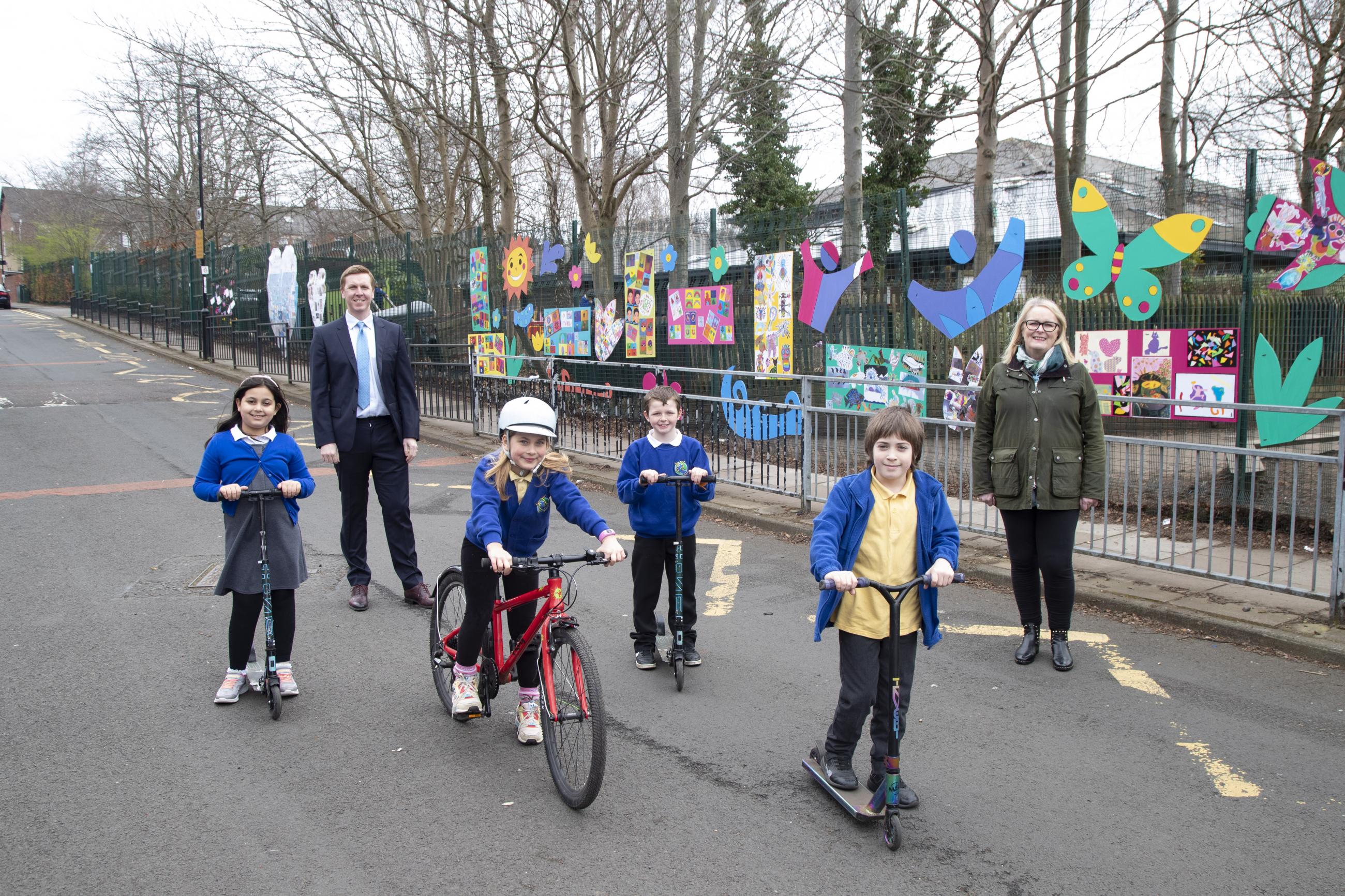 Image showing four primary school children in uniform, three on scooters, one on a bike, outside a school with two adults standing alongside.
