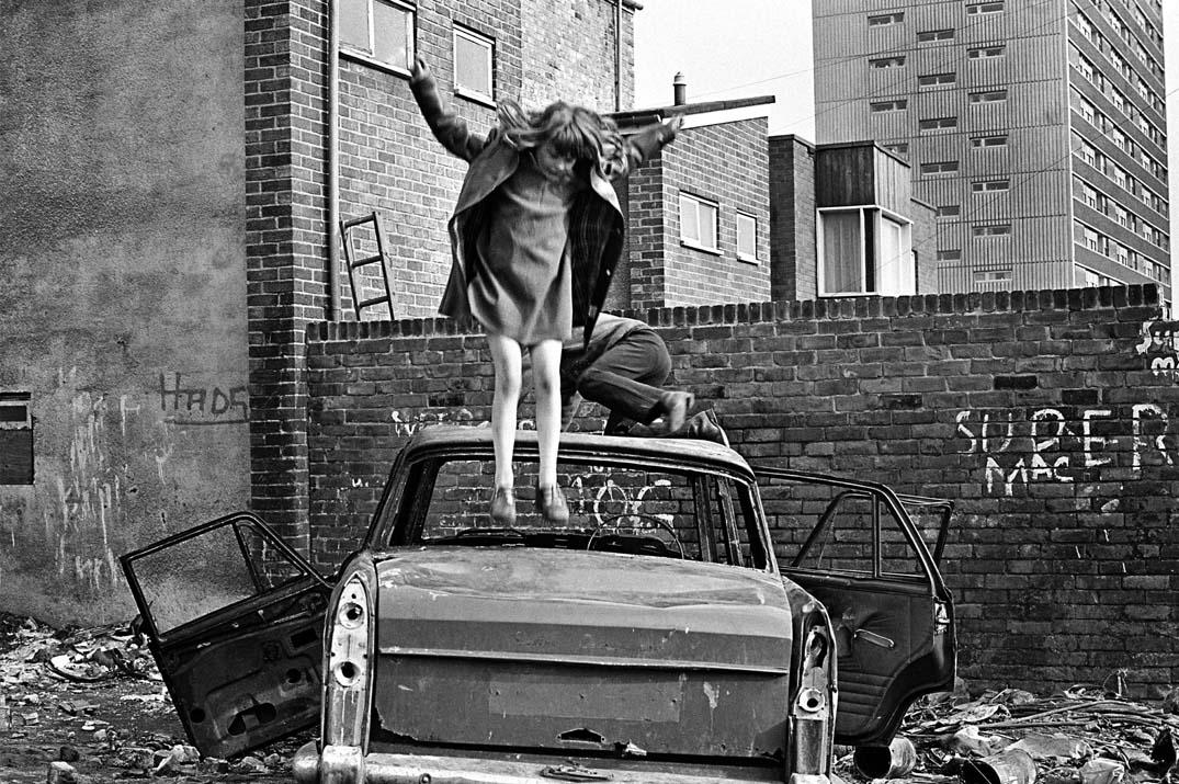 Young girl jumping on a wrecked car with another child climbing behind her. Taken in Elswick in 1978