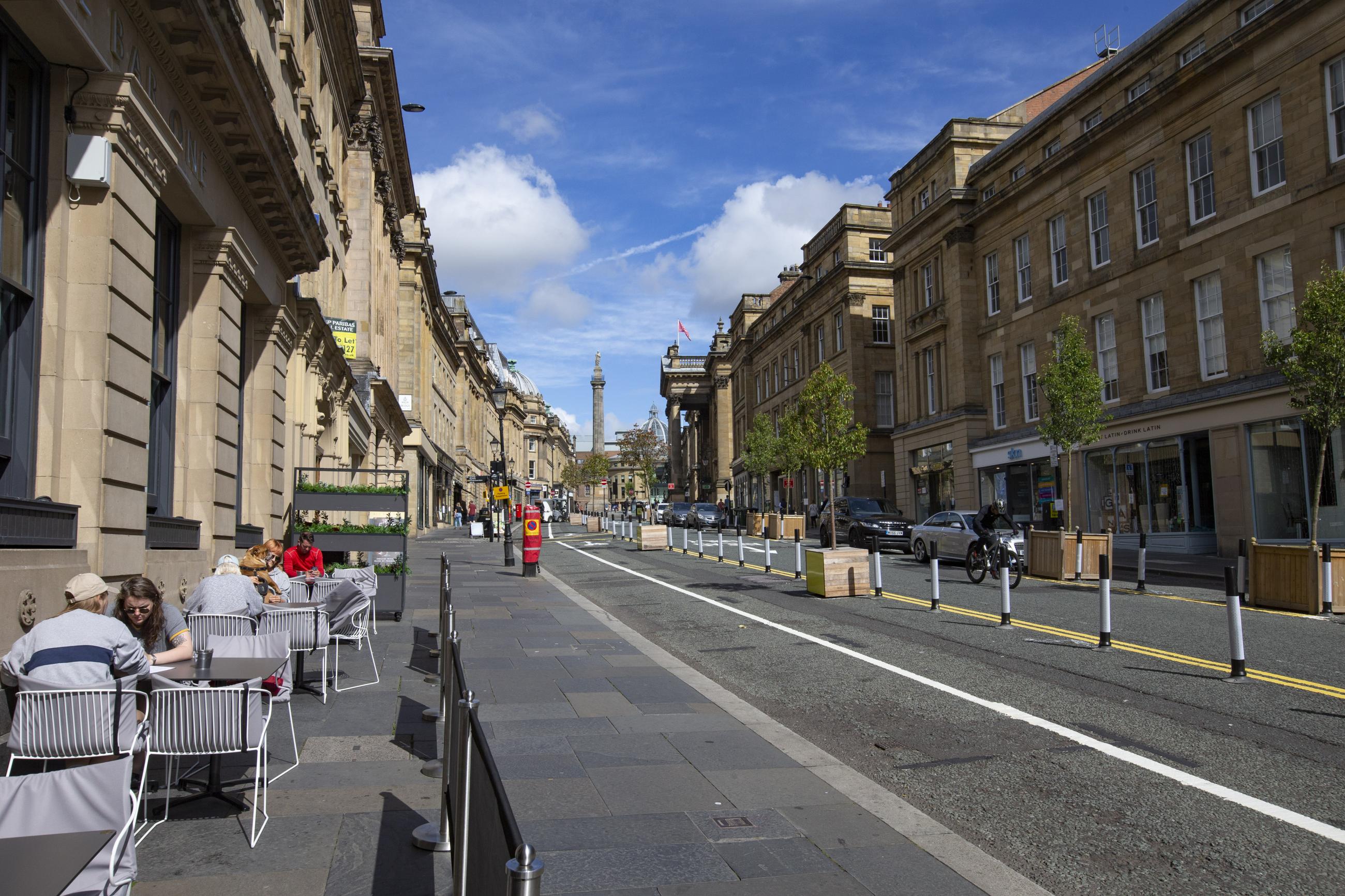 Photo showing Grey Street in Newcastle. There are people sitting at a pavement cafe on the left and part of the road has been sectioned off with temporary bollards to create extra walking space and a cycle lane.