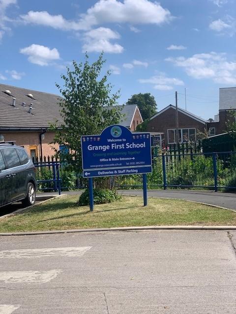 Photo shows the outside of a school with the road in front and a blue sign with the school name in the middle.