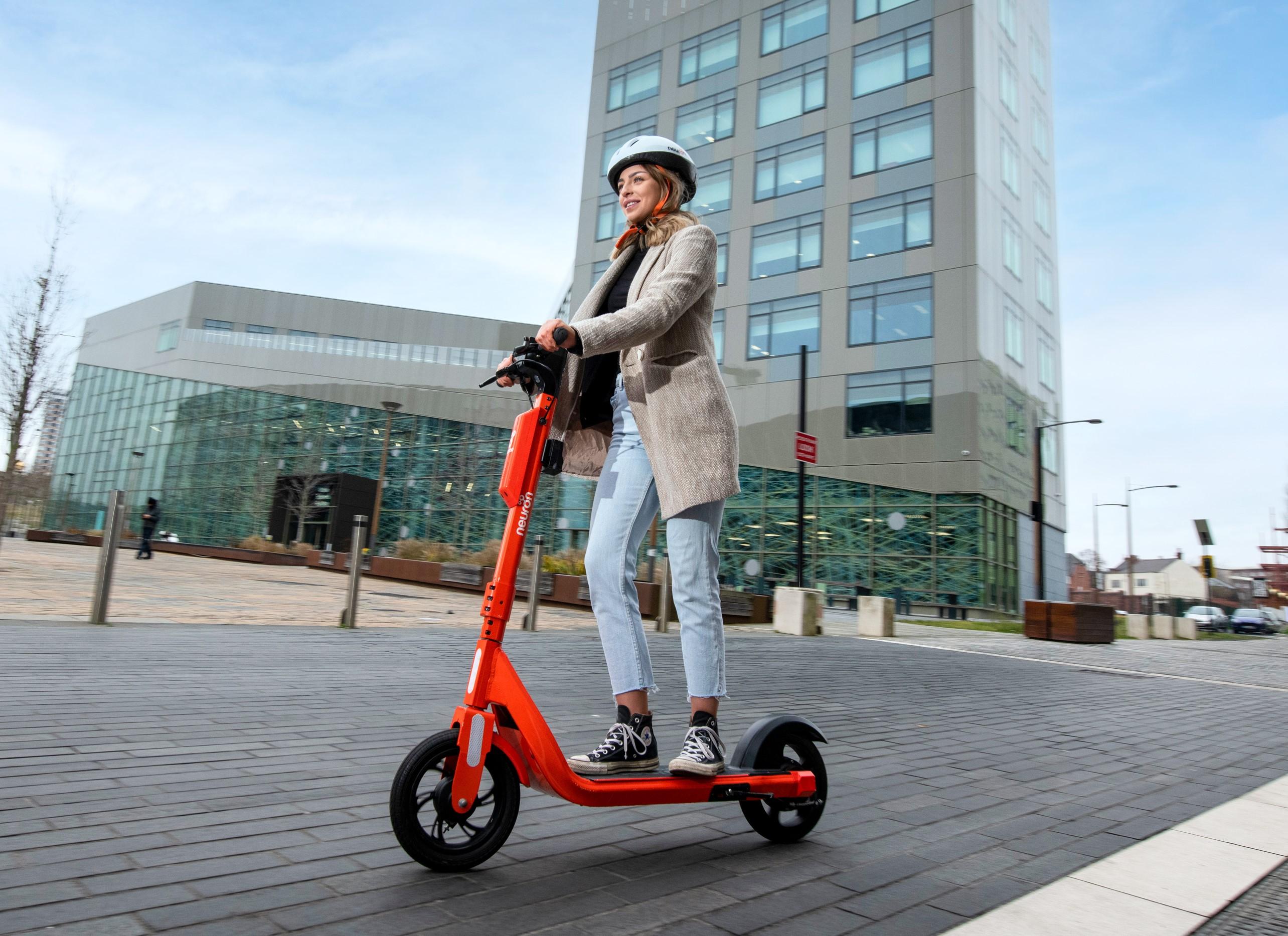 Photo showing a young woman using an orange e-scooter on a clear road with a tall building in the background. She is wearing light coloured clothing and a white helmet.