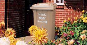 A brown Newcastle City Council garden waste collection bin sat amid colourful flowers, in front of a red brick wall